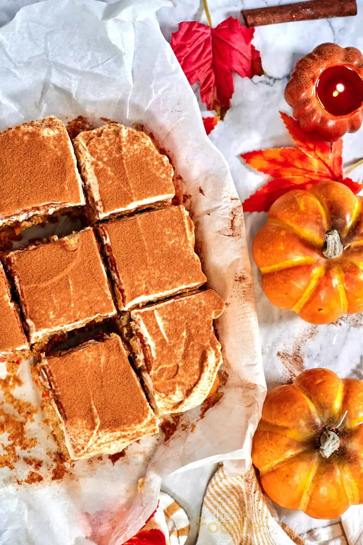 Carrot Cake Bars 11 Flat-lay of cinnamon-topped carrot cake squares beside tiny gourds and a burning pumpkin-shaped candle on a marble surface.