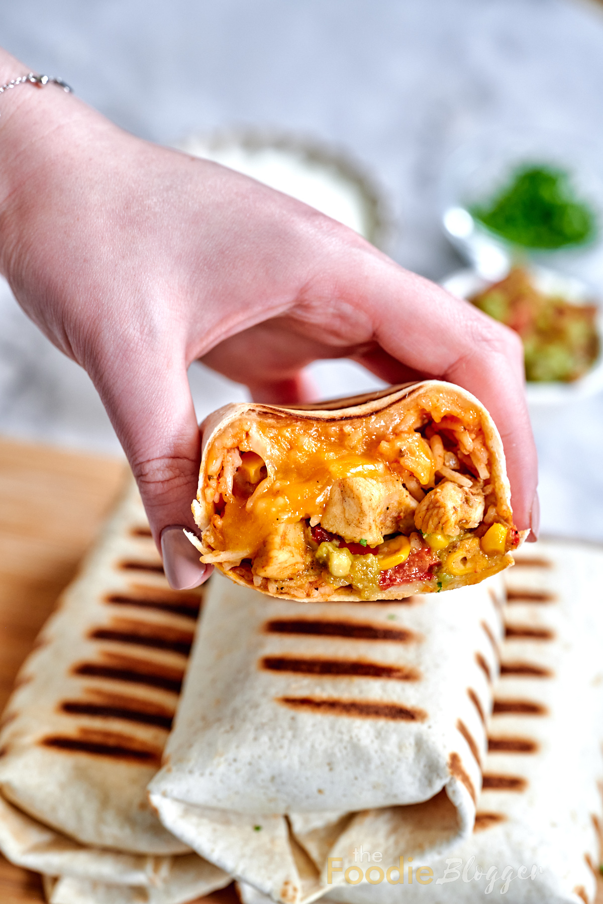 Chicken Burrito 2 A hand lifting a char-marked tortilla half filled with shredded chicken, gooey cheddar, and a smear of green guacamole, with white bowls of condiments behind.
