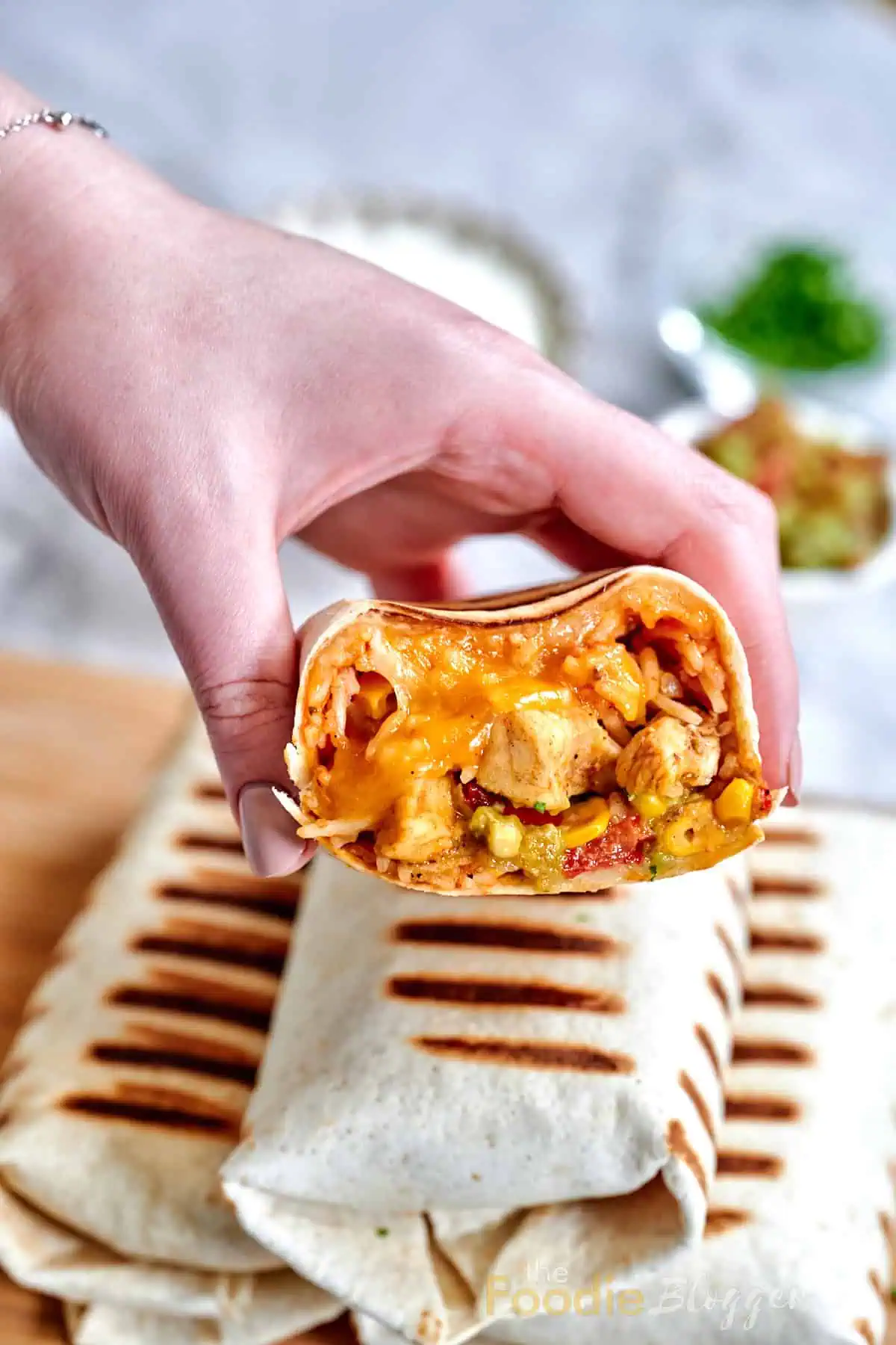 Chicken Burrito 2 A hand lifting a char-marked tortilla half filled with shredded chicken, gooey cheddar, and a smear of green guacamole, with white bowls of condiments behind.