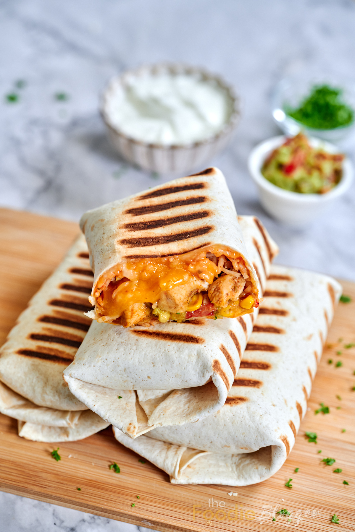 Chicken Burrito 9 Three fully wrapped chicken burritos arranged in a triangular stack on a wood board, the top one sliced open to expose its cheesy, veggie-studded interior.