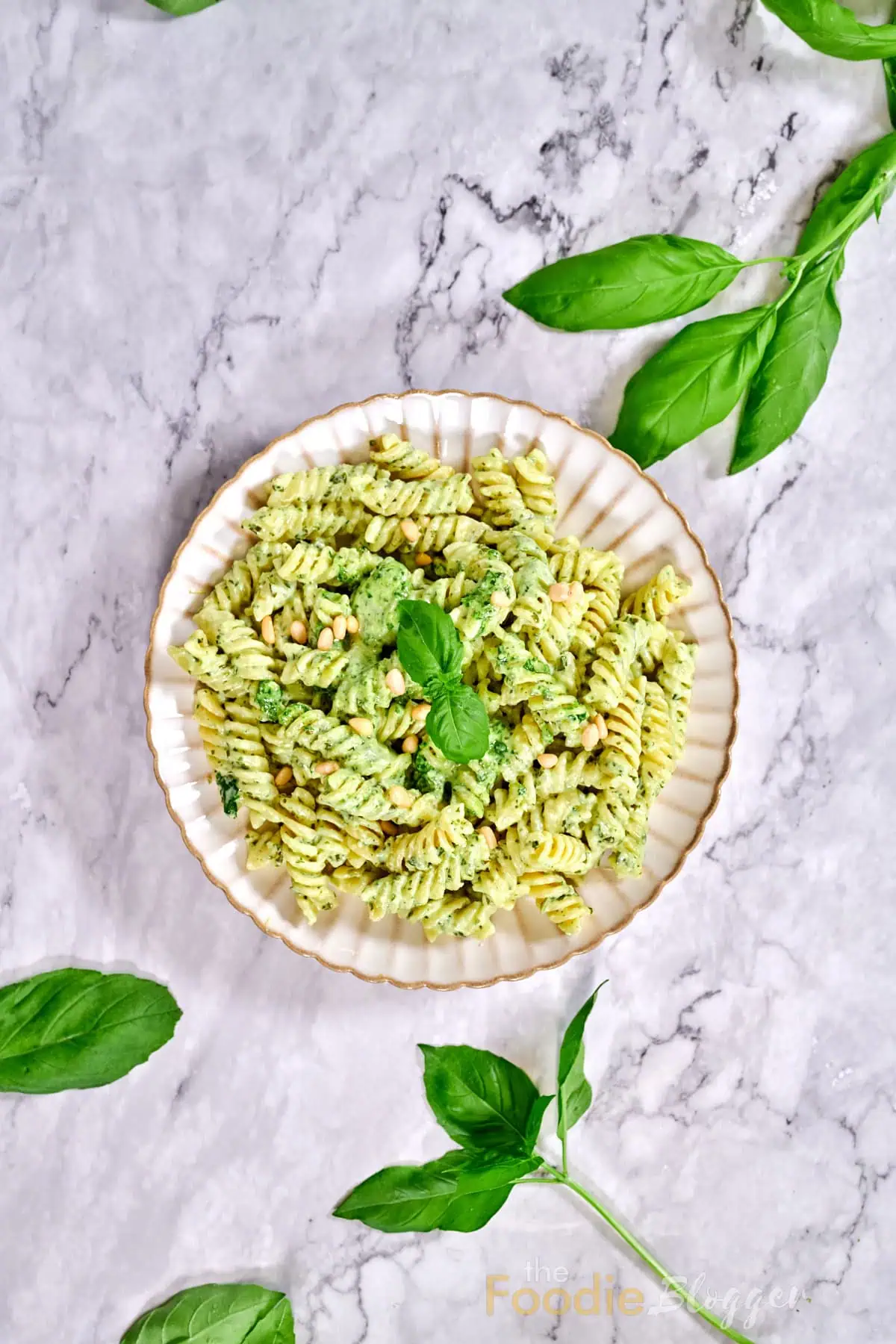 Creamy Pesto Pasta 3 Overhead marble backdrop featuring a centered plate of creamy pesto rotini surrounded by scattered fresh basil sprigs.