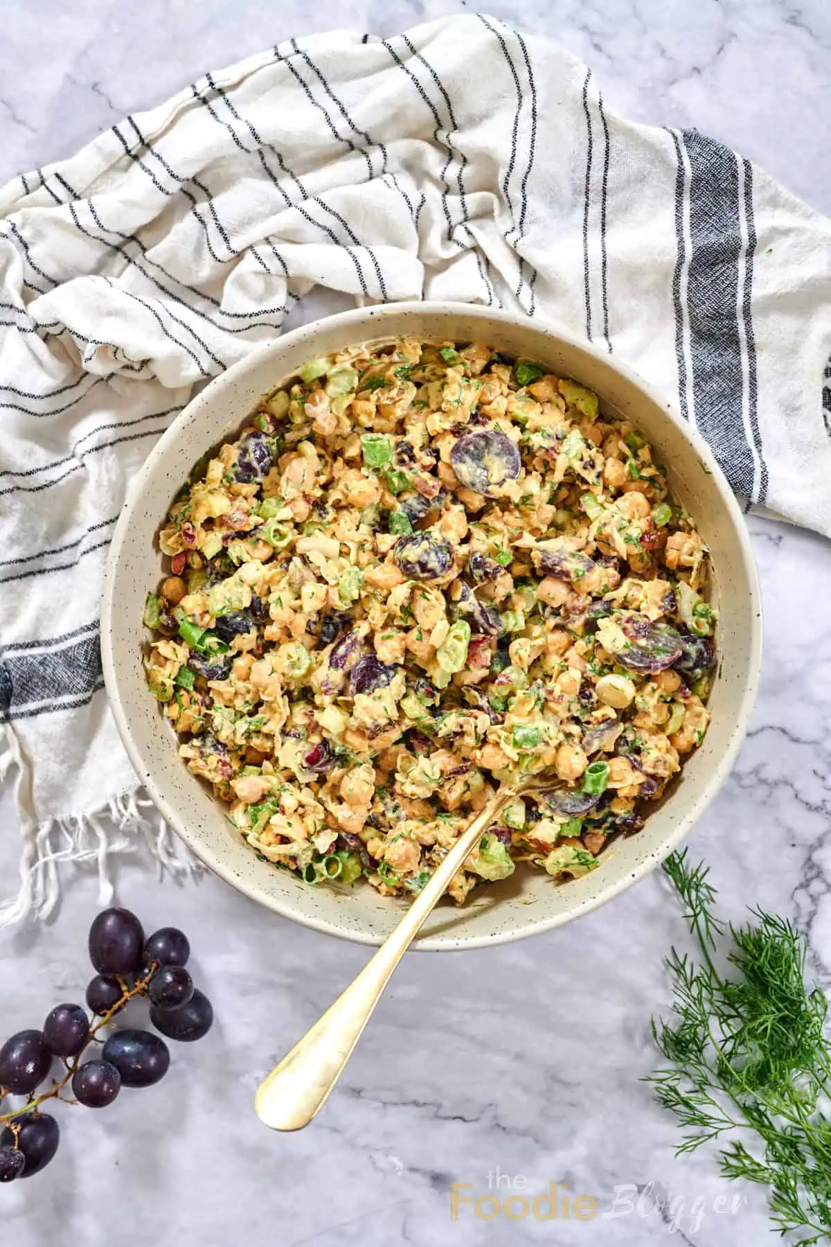 The Foodie Blogger - Tried, True, and Delicious Recipes 12 Overhead view of a large bowl of chickpea salad dotted with grapes, celery, and green onions, spoon ready for serving.
