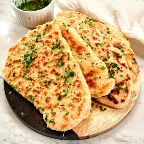 2-Ingredient Garlic Butter Naan 13 Stack of homemade 2-ingredient garlic butter naan brushed with garlic herb butter on a wooden serving board.