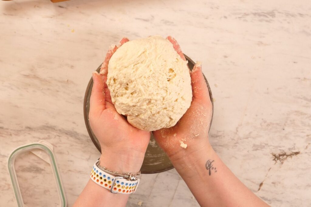 2-Ingredient Garlic Butter Naan 4 Hands holding a soft ball of naan dough over a mixing bowl on a floured countertop.