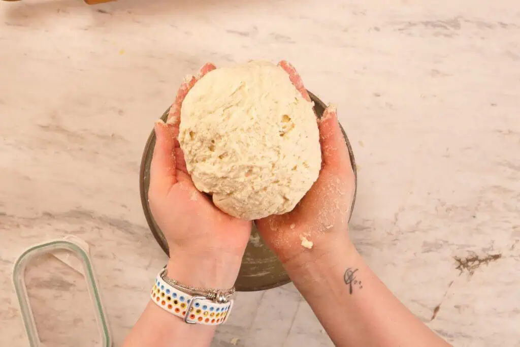2-Ingredient Garlic Butter Naan 4 Hands holding a soft ball of naan dough over a mixing bowl on a floured countertop.